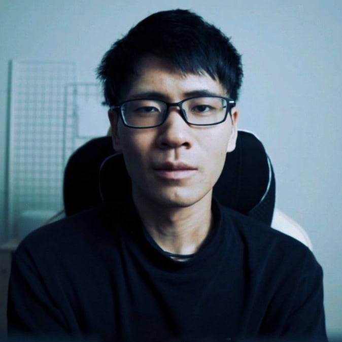 A person with short dark hair and glasses is sitting indoors, looking directly at the camera.